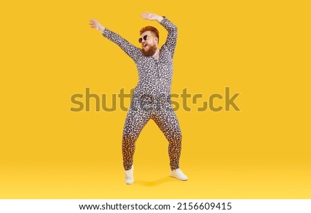 Cheerful funny eccentric fat man in good mood is dancing and fooling around on orange background. Overweight bearded man dressed in leopard print jumpsuit is dancing and fooling around. Full length.