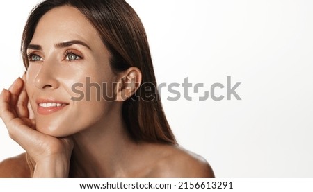 Smiling female aging model, clear glowing skin, nourished face without blemishes, applies daily cream, hyaluron anti-aging treatment, looking in mirror at herself, white background