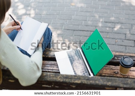 top view of woman writing in notebook near laptop with green screen and paper cup