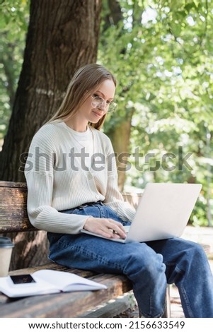 woman in glasses using laptop and sitting on bench in green park