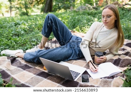 woman in glasses making notes while holding paper cup near laptop and smartphone on blanket in park