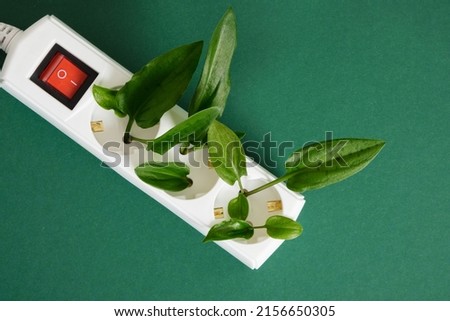 white surge protector with a red power button and green leaves of a plant on a green background, eco-friendly electricity, alternative types of electricity generation
