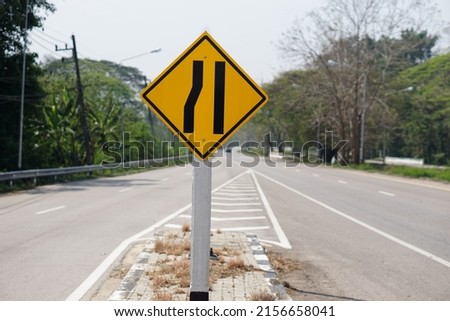 Yellow warning road sign on cement pole at rural street in Thailand. Narrow way traffic symbol. Left narrow lane sign                             