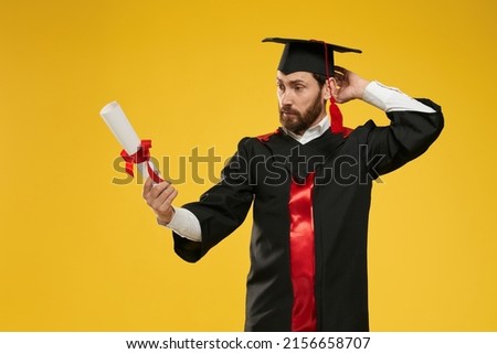 Front view of student standing, looking at diploma, mixed up, surprised. Young male wearing mortarboard and graduate gown graduating from college. Isolated on yellow background.