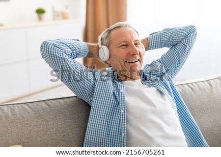Rest And Relax Concept. Excited mature man sitting leaning back on couch listening to music, audio book or podcast, enjoying favorite playlist in wireless headphones holding hands behind head, closeup