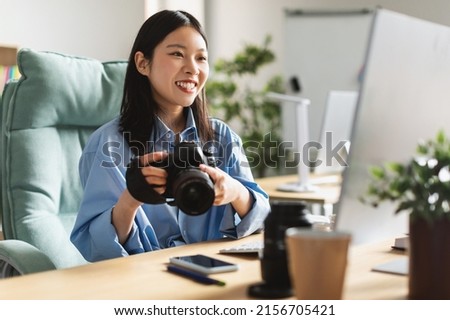 Joyful Professional Asian Photographer Lady Holding Camera Using Laptop Sitting At Workplace, Looking At Screen. Professional Artist Transfer Pictures To Computer For Retouch. Creative Career Concept