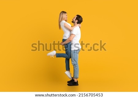 Cheerful young european male with beard raises female, isolated on yellow background, studio, full length, profile. Relationship, love, romance, advertising and offer, win and huge sale, copy space Royalty-Free Stock Photo #2156705453