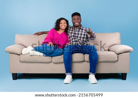 Happy young black couple with remote control sitting on sofa and watching TV over blue studio background, full length. African American spouses with controller enjoying television program or show Royalty-Free Stock Photo #2156705499