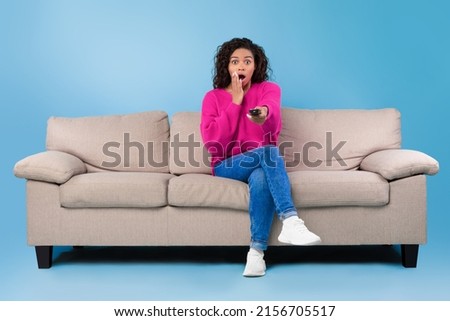 Frightened young African American lady with remote control sitting on sofa and watching scary movie or thriller on TV over blue studio background, copy space. Domestic entertainments concept
