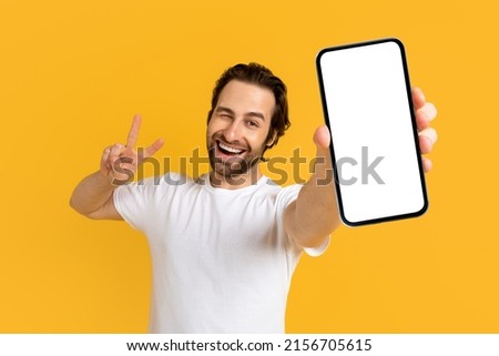 Cheerful emotional young european guy in white t-shirt show phone with empty screen and peace sign with hand isolated on yellow background, studio. Gesture, big sale, new app, device, app and offer Royalty-Free Stock Photo #2156705615