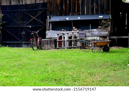 A view of a rural field with some equipment scattered around, including an old bike, a wheelbarrow, some tools, and other farming utiensils seen on a sunny summer day on a Polish countryside
