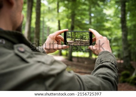 Back view of crop male hiker using smartphone and taking picture of green forest while traveling in summer day