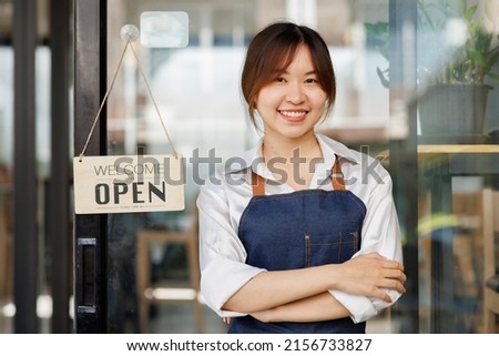 Portrait of a positive Asian businesswoman standing at the cafeteria door entrance. A cheerful young waitress in a blue apron near a glass door with an open signboard.SME Excited small business owner.