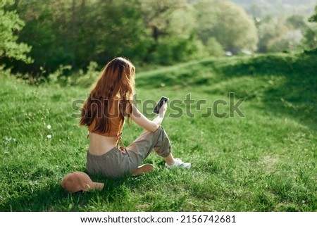 Top view of a woman in an orange top and green pants sitting on the summer green grass with her back to the camera with her phone, a young freelance student's concept of work and leisure Royalty-Free Stock Photo #2156742681