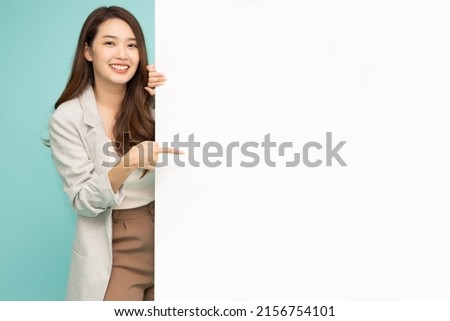 Cheerful Young Asian businesswoman is standing behind the white blank banner or empty copy space advertisement board on green background, Looking at camera