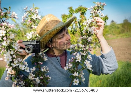 Photography as an interesting hobby. Middle-aged woman in a straw hat holding a vintage camera among the branches of a blossoming apple tree.