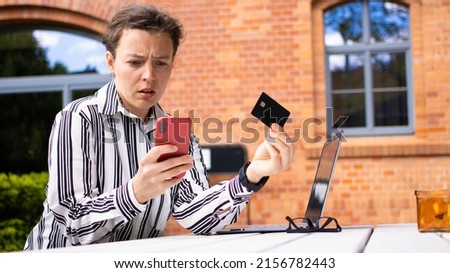Confused business woman holding credit card, looking at mobile phone sitting at table outdoors red brick building background. Debt problems, failed transaction, money error, contacting with scammer.