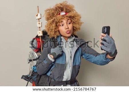 Active tourist takes selfie photo keeps lips folded dressed in sportswear carries heavy rucksack leads active lifestyle has journey expedition isolated over grey wall. Female hiker returns from picnic