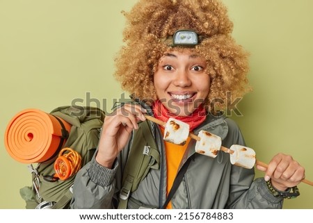 Happy curly haired woman camper holds wooden stick with roasted marshmallow wears headlamp and windbreaker carries rucksack isolated over green background. Young tourist has picnic during weekend