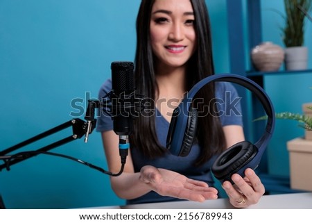 Asian vlogger creator holding headset talking into studio microphone with subscribes recording headphones review. Social media influencer filming tutorial using professional equipment in studio