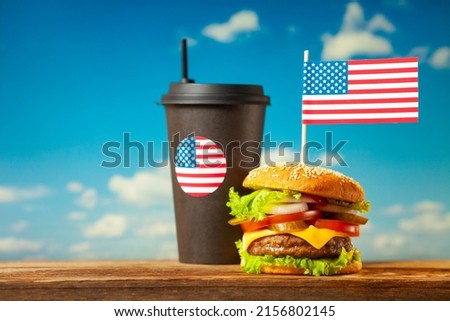 Close-up home made beef burger with american flag on the top and black paper cup on wooden table over blue sky background.