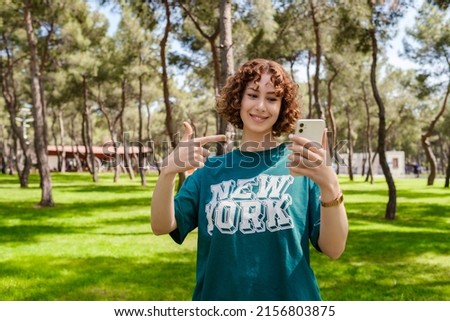 Redhead woman pointing to phone with big smiles while standing on a city park. Woman wearing green tee and golden wrist watch. Sportive woman using phone concepts.