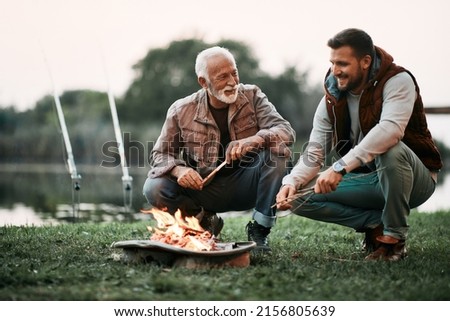 Happy mature man and his adult son talking while relaxing by campfire during their fishing day. Royalty-Free Stock Photo #2156805639
