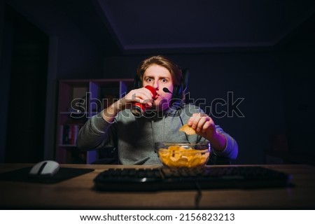 Emotional guy gamer with a shocked face playing online games on the computer, drinking soda and eating chips from a plate at home in a dark room