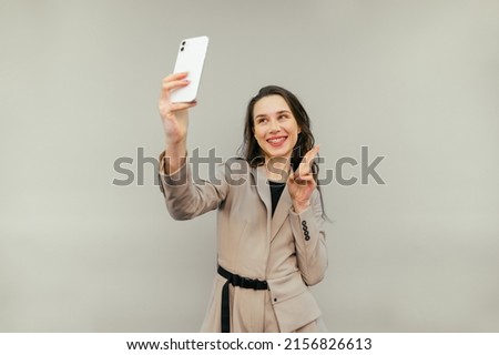 Joyful lady in a jacket stands on a beige background with a smile on her face takes a selfie and shows a gesture of peace, posing in the smartphone camera.