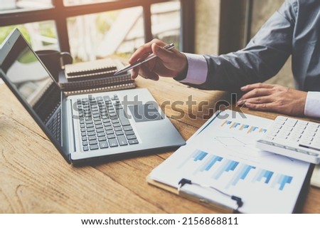 Business man hold pen and pointing to laptop at office with document report, graph, financial report on screen. Royalty-Free Stock Photo #2156868811