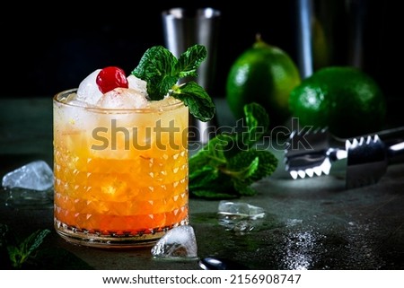 Mai Tai popular alcoholic cocktail with rum, liqueur, syrup, lime juice, mint and crushed ice. Dark green background, steel bar tools, negative space Royalty-Free Stock Photo #2156908747