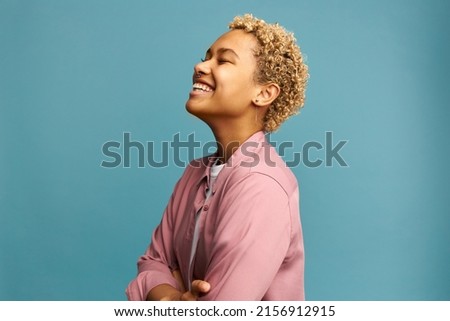 Profile view of happy joyful carefree female with dark skin and blonde curly hair, standing against blue background in pink shirt with arms folded, putting head up. Human emotions and feelings concept Royalty-Free Stock Photo #2156912915