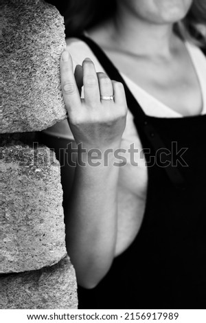 A black and white photograph of a woman standing against a wall. The hand with the ring is in focus. Vertical photo.