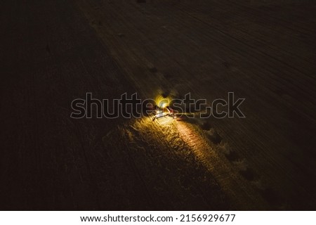 An aerial drone shot of a harvester machine working on a field with lights on at night