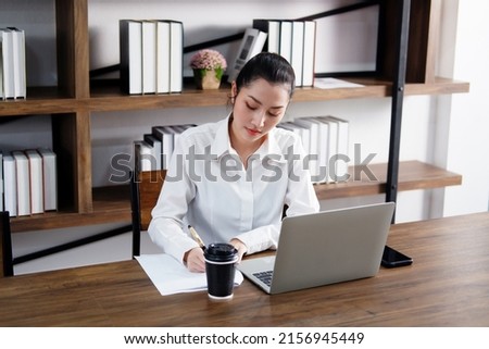 Asian businesswoman is online working with laptop and writing information on paper in workspace at office. Collegian woman are researching information on the Internet in library at the university. Royalty-Free Stock Photo #2156945449