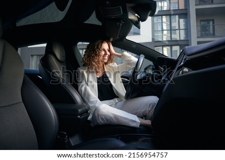 Side view from inside car. Business lady resting in the driver's seat of comfortable elegance automobile, smiling happily and looking to the right. On the background residential building.