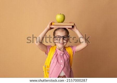 Back to school kid. Happy blonde child holds a books and a green apple in her head on a beige background. Education and intellectual development of children. World book day