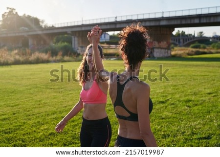 Stock photo of slender girls in sportswear holding hands on green field at sunset, bridges on background. Best friends cheering up and training together outside. Concept of fitness