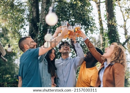 happy young friends toasting with beer together - cheerful people clinking glasses outdoor at party - multiethnic group having fun and celebrating