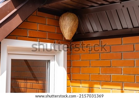 Life hack. Wasp nest decoy of paper in form of elongated ball under roof of country house. Close-up of false wasp nest under brown metal profile roof. Brick wall made of orange Italian facing bricks. Royalty-Free Stock Photo #2157031369