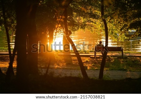Silhouette of a lonely man standing alone on a bench from a park during a summer sunset.