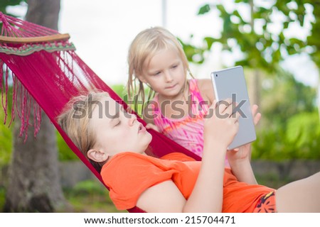 Young woman show pictures to adorable daughter on electronic tablet reader relaxing in hammock under palm trees