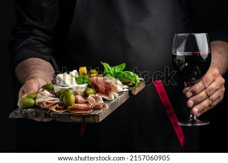 Hands holding a glass of wine and a wooden board with different kinds of cheese and ham, prosciutto, jamon salami, Antipasto Dinner or aperitivo party. Royalty-Free Stock Photo #2157060905