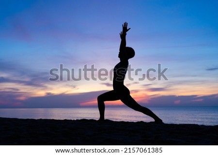 Silhouette of a beautiful woman practicing yoga asana on the beach with sunrise on twilight blue vibrant sky and calm sea in background. High lunge, Ashta Chandrasana, June 21, International Yoga Day. Royalty-Free Stock Photo #2157061385
