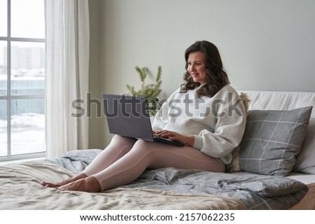 young beautiful pregnant woman sits on the bed in cozy clothes and looks at something on the laptop and u. tender photo. concept of future motherhood