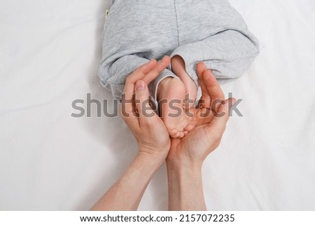 Mom's hands gently hold the baby's legs in gray pants on a white background, space for text Royalty-Free Stock Photo #2157072235