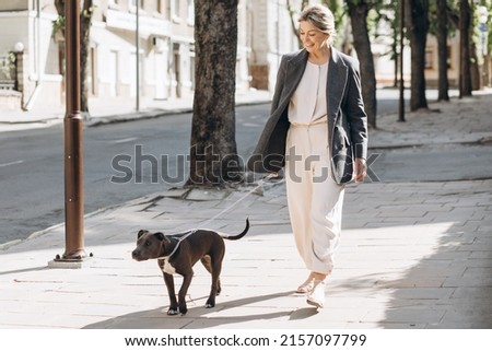 Mature blond business woman in a gray jacket smiling walking her dog breed amstaff and talking on the phone