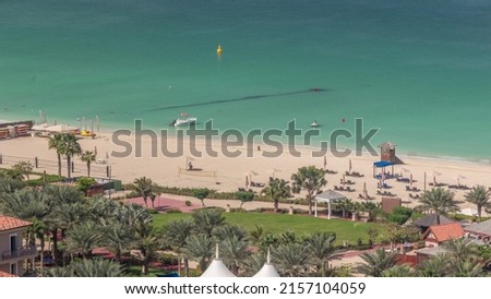 The beach at JBR with golden sand near seaside aerial timelapse. Green lawn with palms and people relaxing on chairs under umbrellas with motor boats on a water