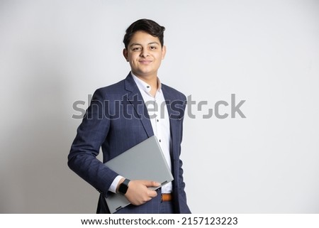 Portrait of young confident Indian teenager boy wearing suit holding laptop in hand looking at camera isolated on white studio background. Royalty-Free Stock Photo #2157123223