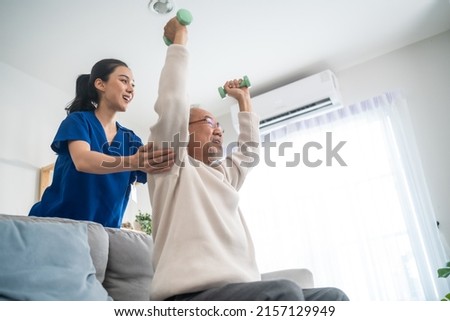Asian older aged man doing physiotherapist with support from nurse. Senior elderly male sitting on sofa in living room using dumbbells workout exercise for patient with caregiver in nursing care. Royalty-Free Stock Photo #2157129949